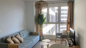 Un salon moderne avec un canapé gris, une table à manger avec des chaises, une grande fenêtre avec des rideaux et un luminaire suspendu.