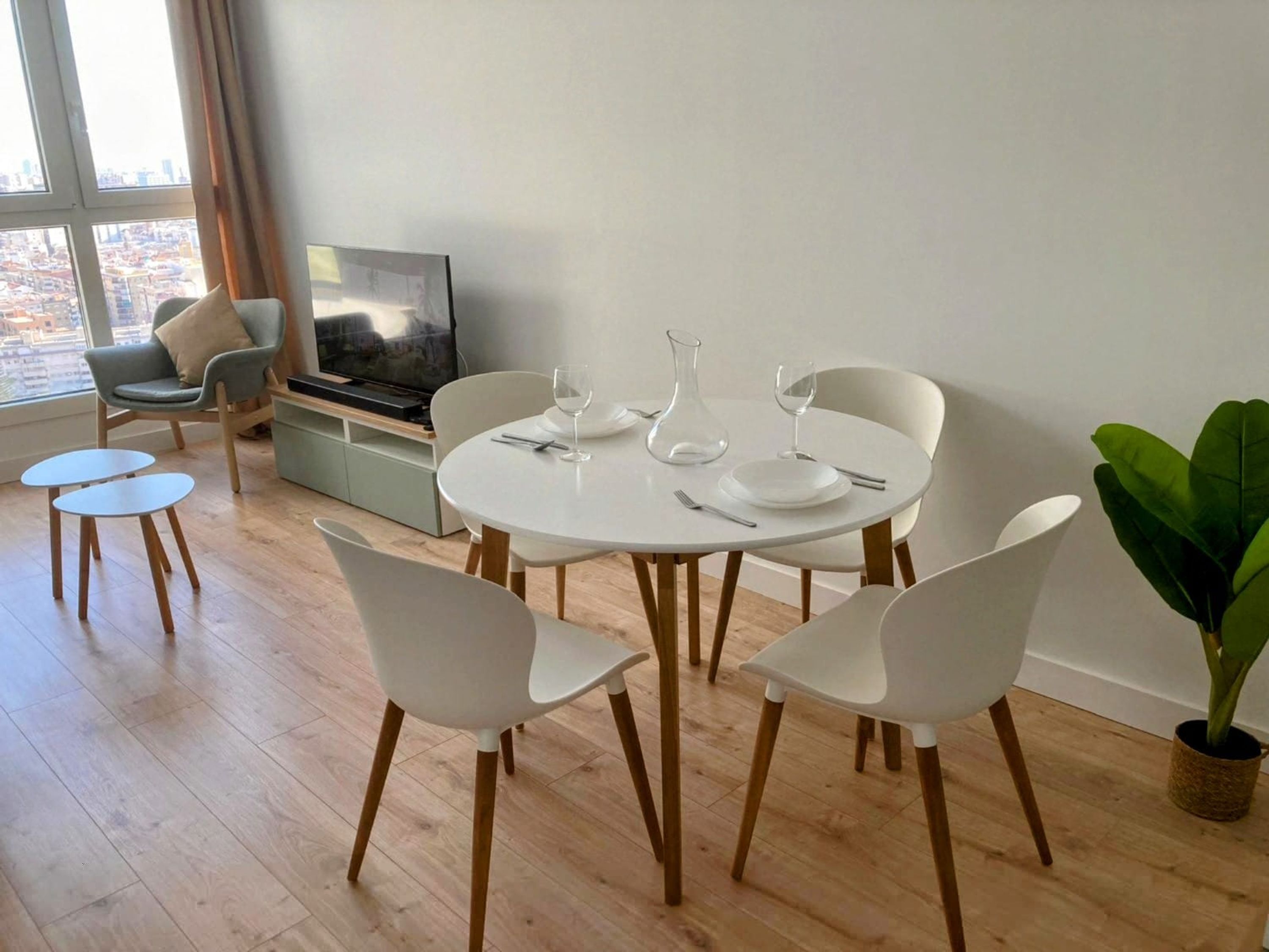 A modern dining area with a round table set for four, surrounded by white chairs. A television is mounted on the wall, and a large window offers a view of the cityscape.
