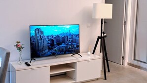 A modern living room with a flat-screen TV on a white entertainment unit, a standing lamp, a clock on the wall, and a fluffy rug on the floor. - Happy Travel La Maestranza 5A (Marbella)