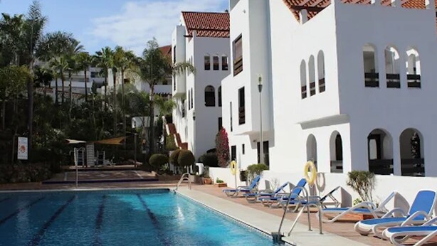 A swimming pool area with lounge chairs and a building with a tiled roof. - Happy Travel La Maestranza 5A (Marbella)