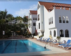 A swimming pool area with lounge chairs and a building with a tiled roof. - Happy Travel La Maestranza 5A (Marbella)