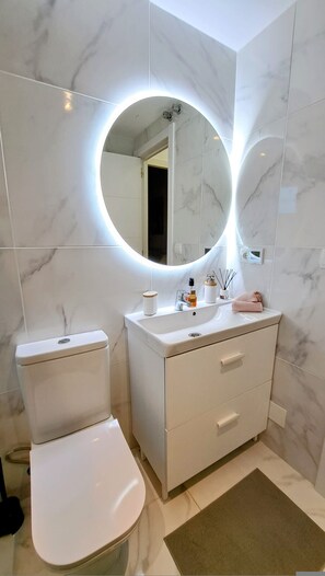 A modern bathroom with a toilet, vanity, and a round mirror with built-in lighting. - Happy Travel La Maestranza 5A (Marbella)
