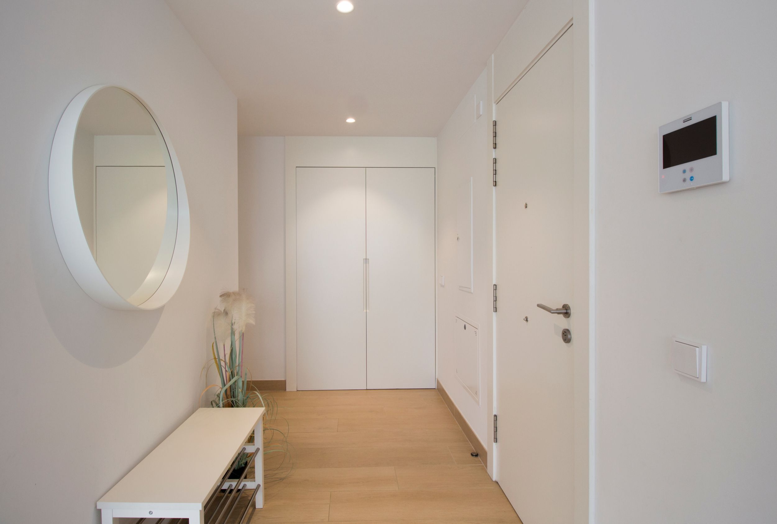 A hallway with a white bench, a round mirror, a door with a digital lock, and a thermostat on the wall.