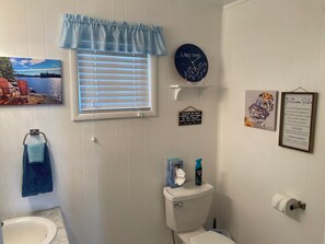 A bathroom with a toilet, a sink, a window with blinds, a wall clock, and a framed sign.