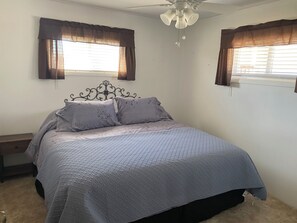 A bedroom with a bed, two windows with curtains, a ceiling fan, and a nightstand.