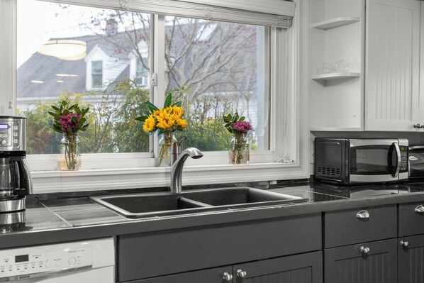 A modern kitchen with a stainless steel sink, a Bosch dishwasher, and a microwave. There are three flower vases on the windowsill.