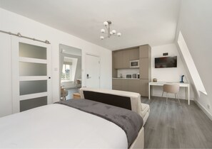 A modern bedroom with a large bed, a desk, and a kitchenette. - YTD Residential (London)