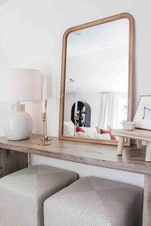 A room with a large mirror, a lamp, and a small table with a picture frame. There are two cushioned seating areas.