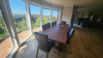 Een ruime eetruimte met een grote houten tafel en moderne stoelen, grote ramen met uitzicht op een bergachtig landschap, en een kroonluchter aan het plafond.