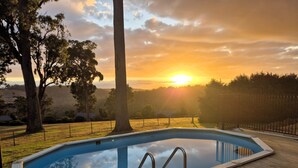 Ein rechteckiger Pool mit Holzterrasse, umgeben von Bäumen und einem Zaun, mit Sonnenuntergang im Hintergrund.