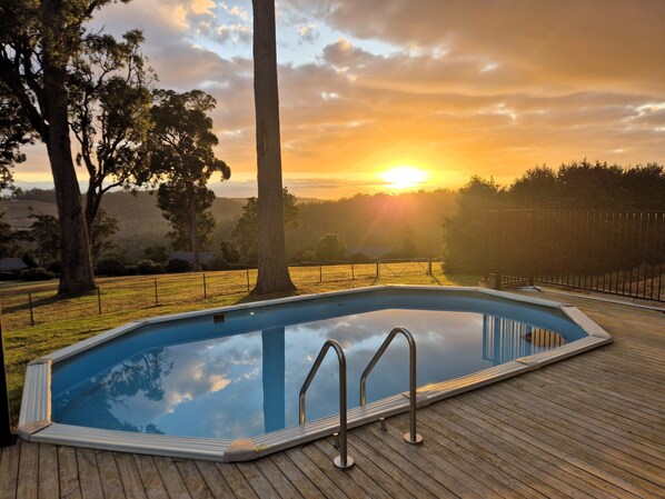 Piscina rectangular con plataforma de madera, rodeada de árboles y una valla, con un atardecer al fondo.