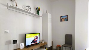 Un salón moderno con un televisor de pantalla plana sobre un soporte de madera, una pared blanca, un sillón individual y una pequeña mesa con una lámpara y un letrero.