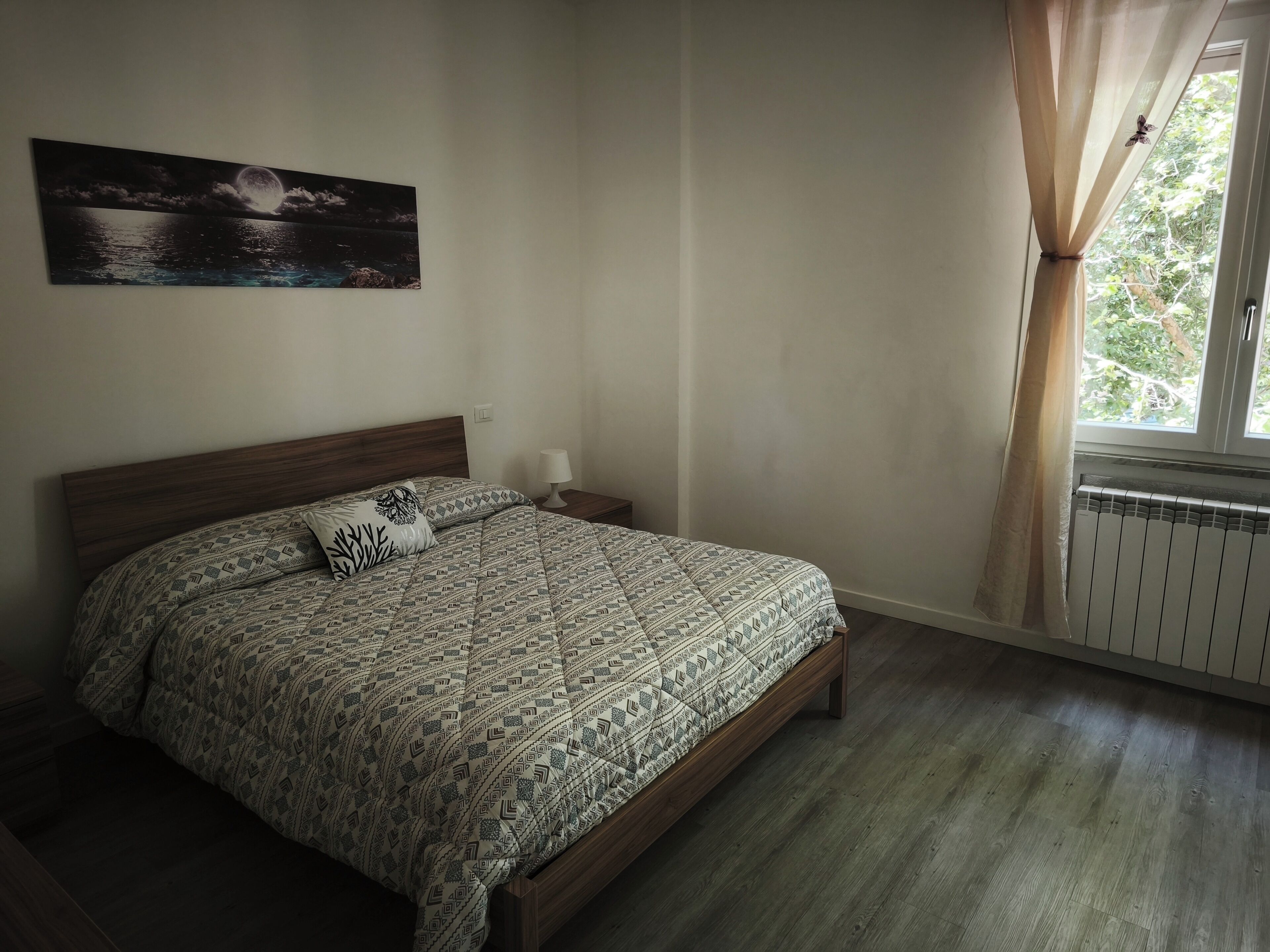 A bedroom with a bed, a nightstand, a wall-mounted lamp, a window with curtains, and a painting on the wall.