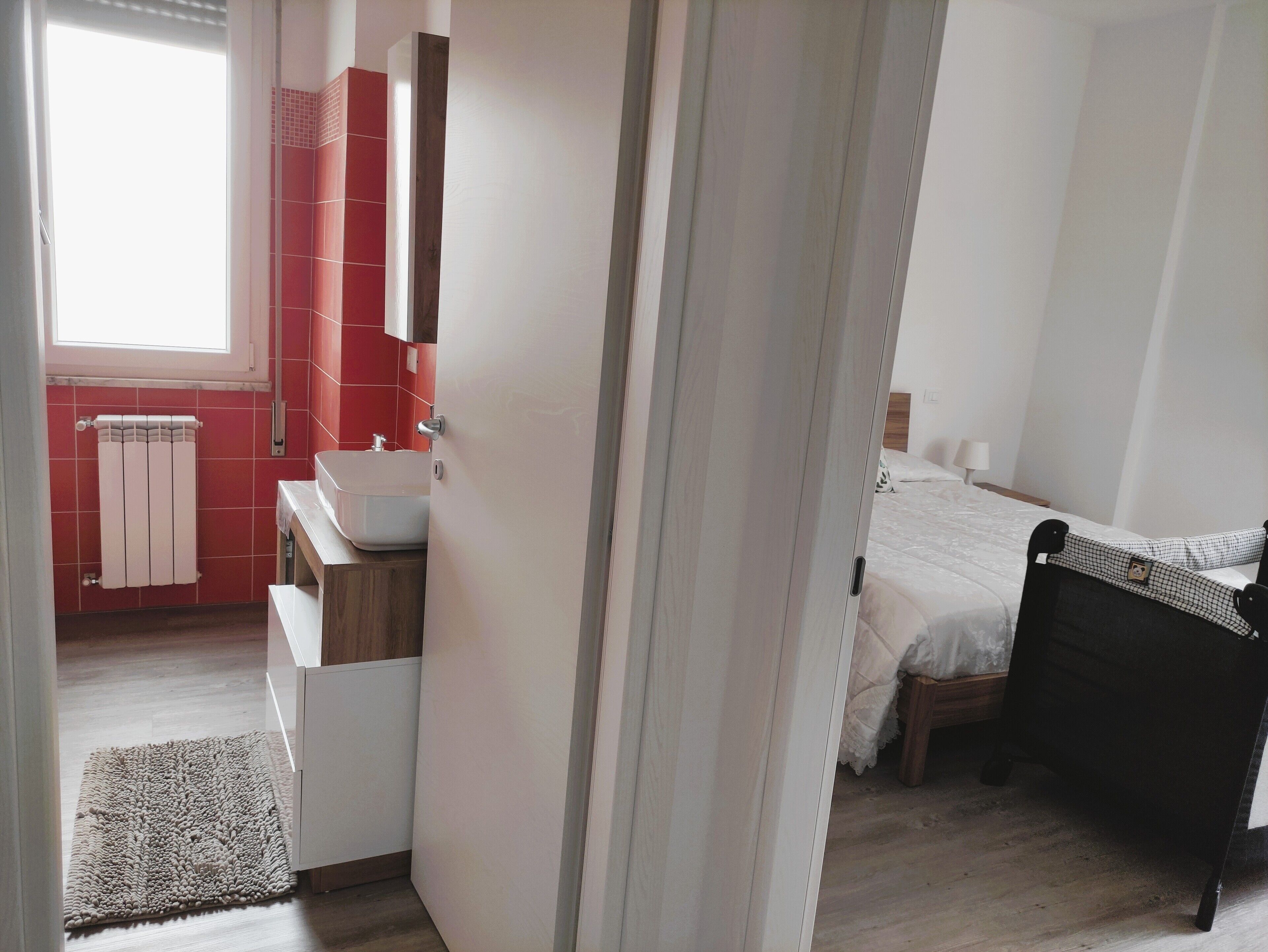 Een hotelkamer met een houten vloer, een wit bed, een zwart hoofdbord, een witte nachtkastje, een witte wand, een raam, een rode betegelde muur, een witte radiator, een witte wastafel, een witte kast, een witte deur en een wit gordijn.