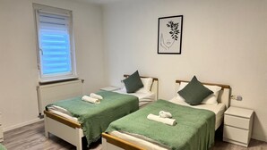 A room with two single beds, each with a green blanket and a rolled towel. There is a window with blinds, a radiator, and a framed picture on the wall. - Apartment in the center with garden, connection to Stuttgart, Märklineum and EWS Arena. (Göppingen)