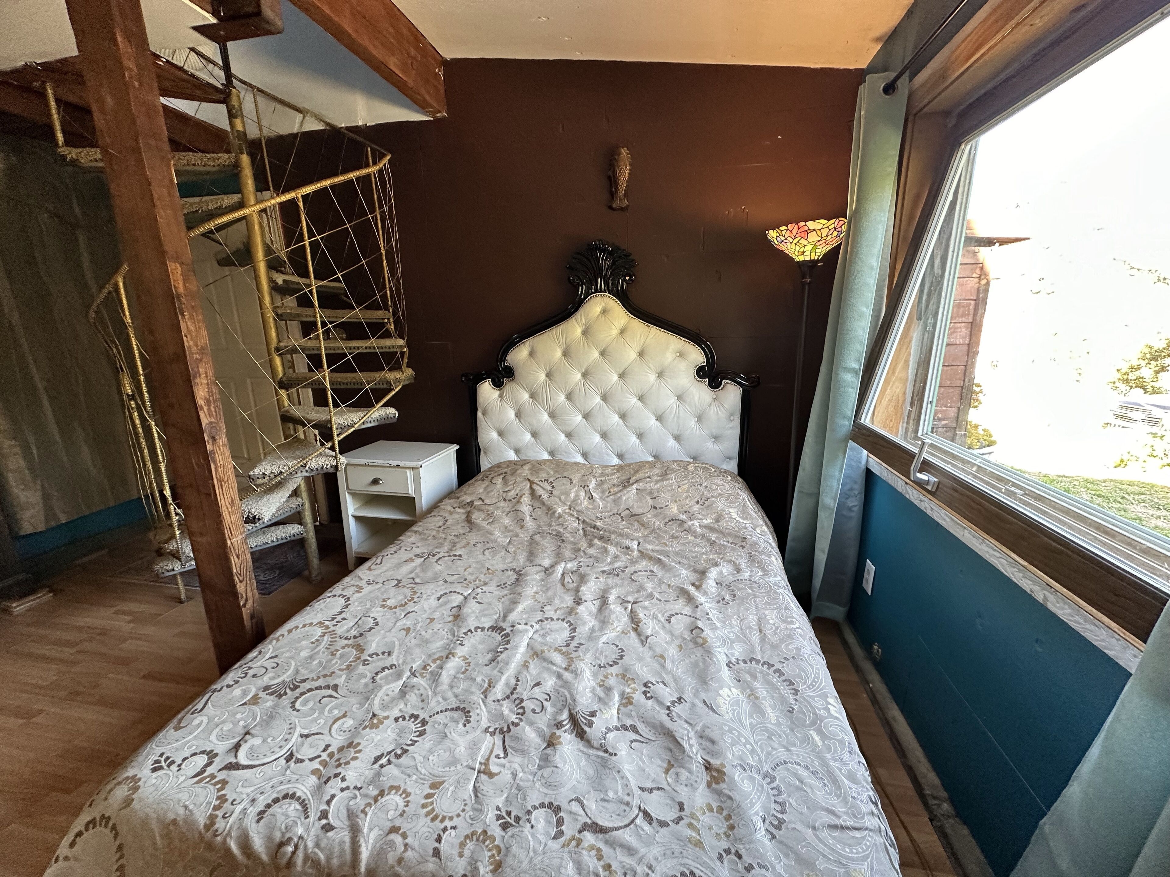 A bedroom with a bed, a small bedside table, a hanging chair, and a window with a view of the outside.