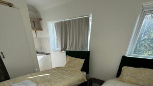 Ein Zimmer mit zwei Betten, jeweils mit floralem Überwurf und weißem Handtuch. Es gibt ein Fenster mit Vorhängen, einen kleinen Tisch und eine Küchenzeile im Hintergrund.