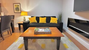 A living room with a black sofa, yellow cushions, a wooden coffee table, a TV mounted on the wall, and a patterned rug.