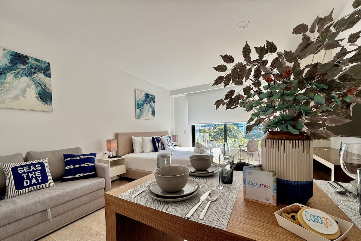 A modern hotel room with a dining area, a sofa with decorative pillows, a large window, and a potted plant.