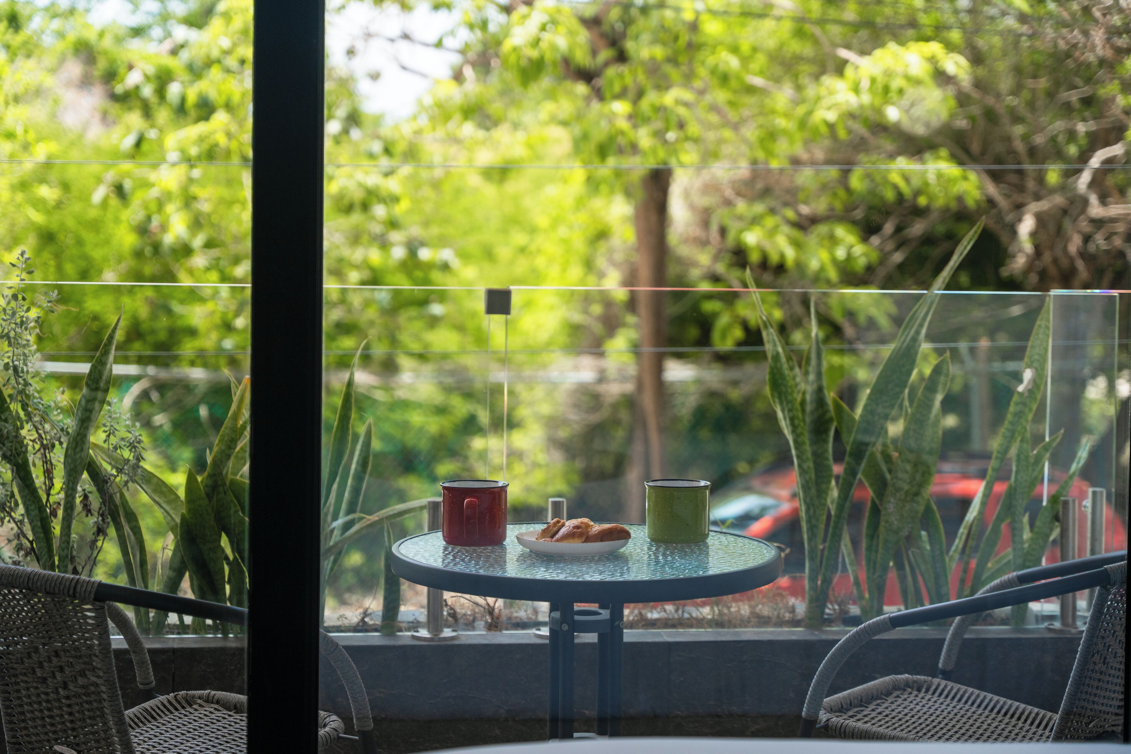 Ein runder Glastisch mit einer Platte Croissants und zwei Tassen, umgeben von Korbstühlen, auf einem Balkon mit Blick auf Grünflächen und parkende Autos.