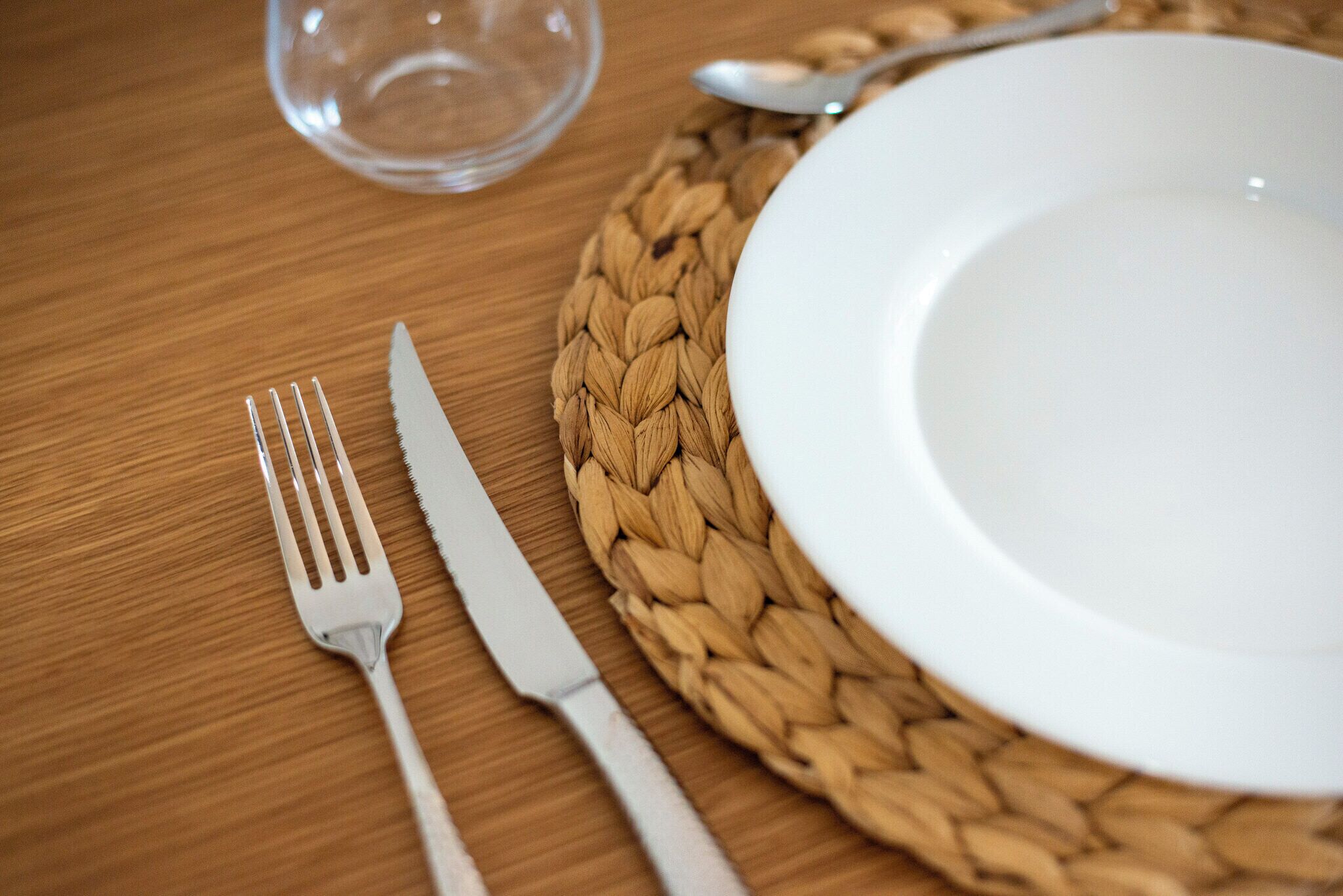 A table setting with a white plate, a fork, a knife, and a spoon on a woven placemat.