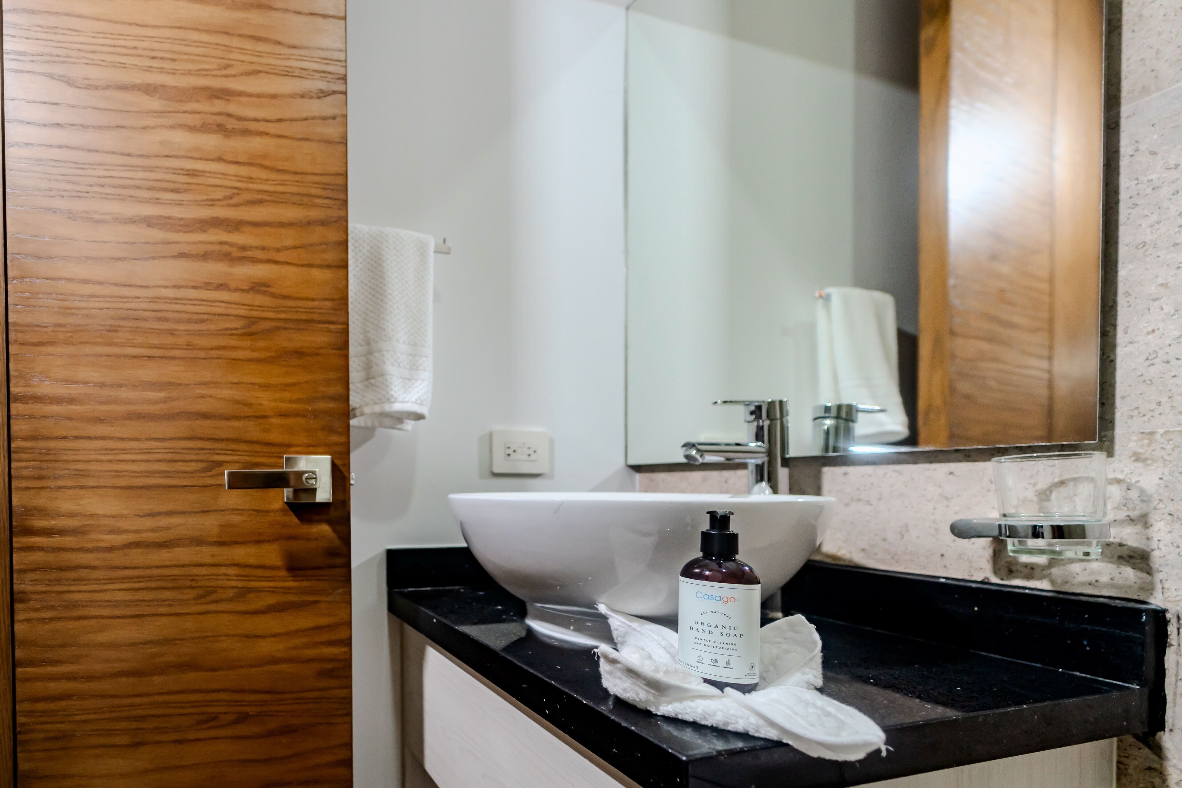 A bathroom with a wooden door, a white sink, a mirror, and a bottle of organic hand soap.