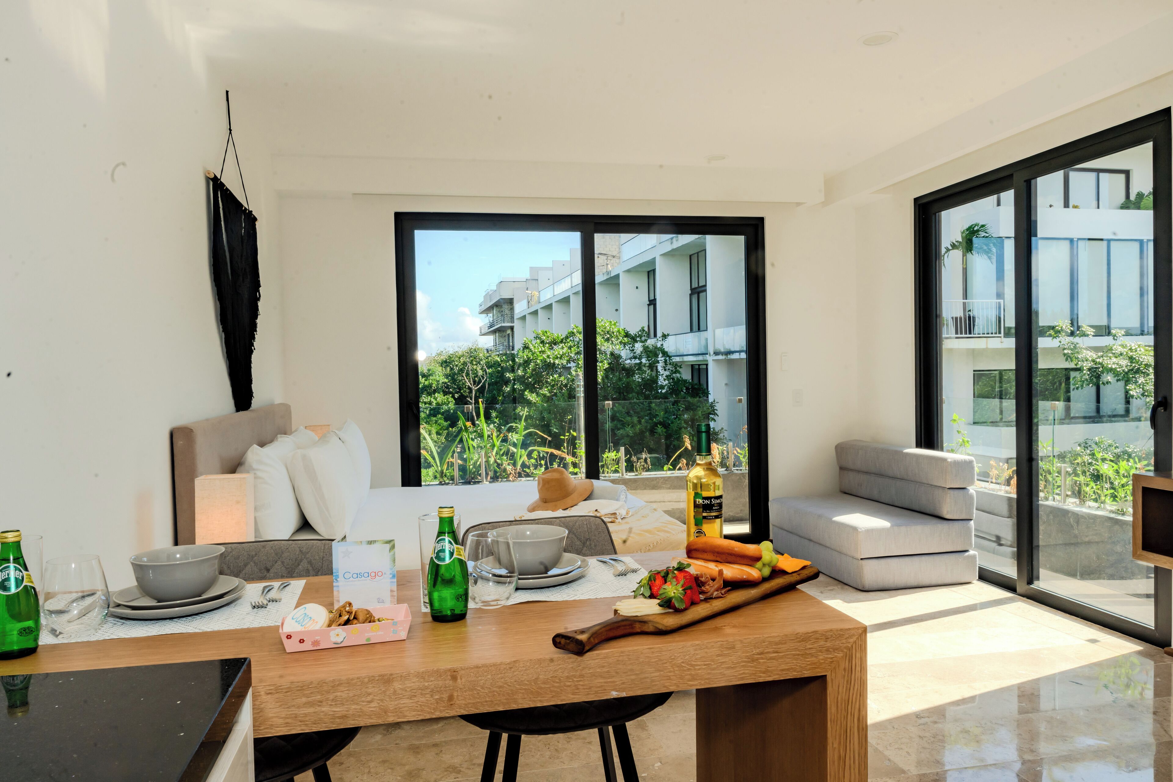 Un salón moderno con un sofá, una mesa de comedor con platos, vasos y una botella. Hay un ventanal con vistas a edificios y zonas verdes.