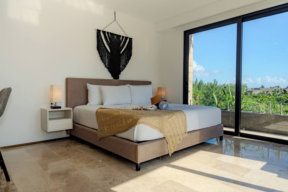 A modern bedroom with a large bed, bedside tables, a hanging macrame wall decoration, and a view of greenery through large windows.