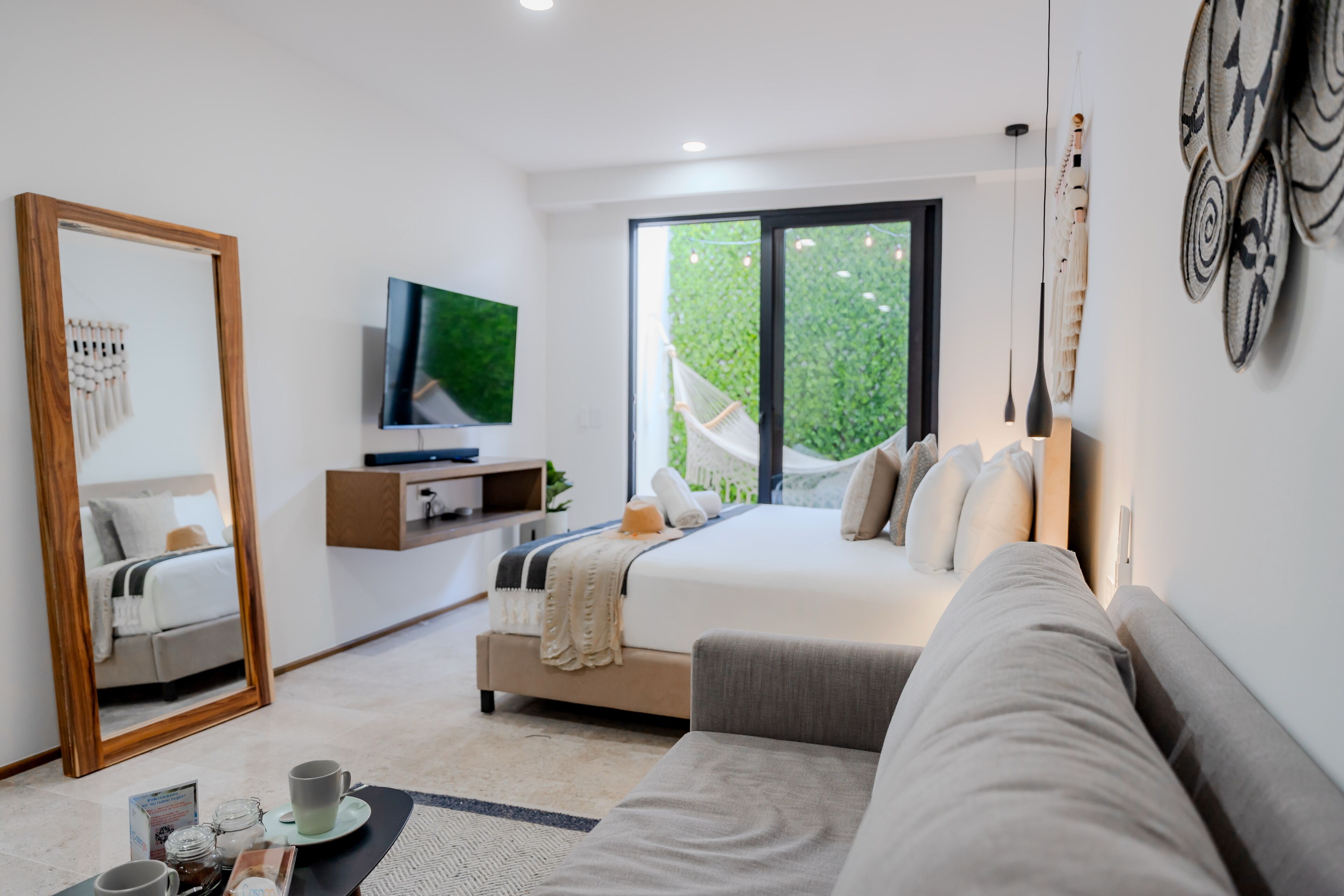Ein modernes Schlafzimmer mit einem großen Bett, einer Couch, einem Fernseher und einem großen Fenster mit Blick auf Grünflächen.