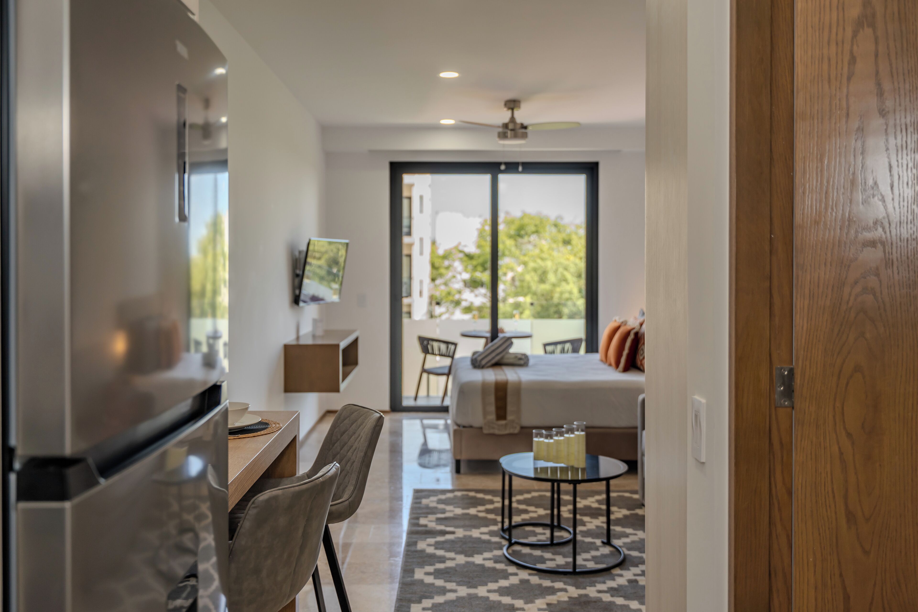 Una habitación de hotel moderna con kitchenette, zona de comedor, cama con vistas y una pequeña mesa con vasos.