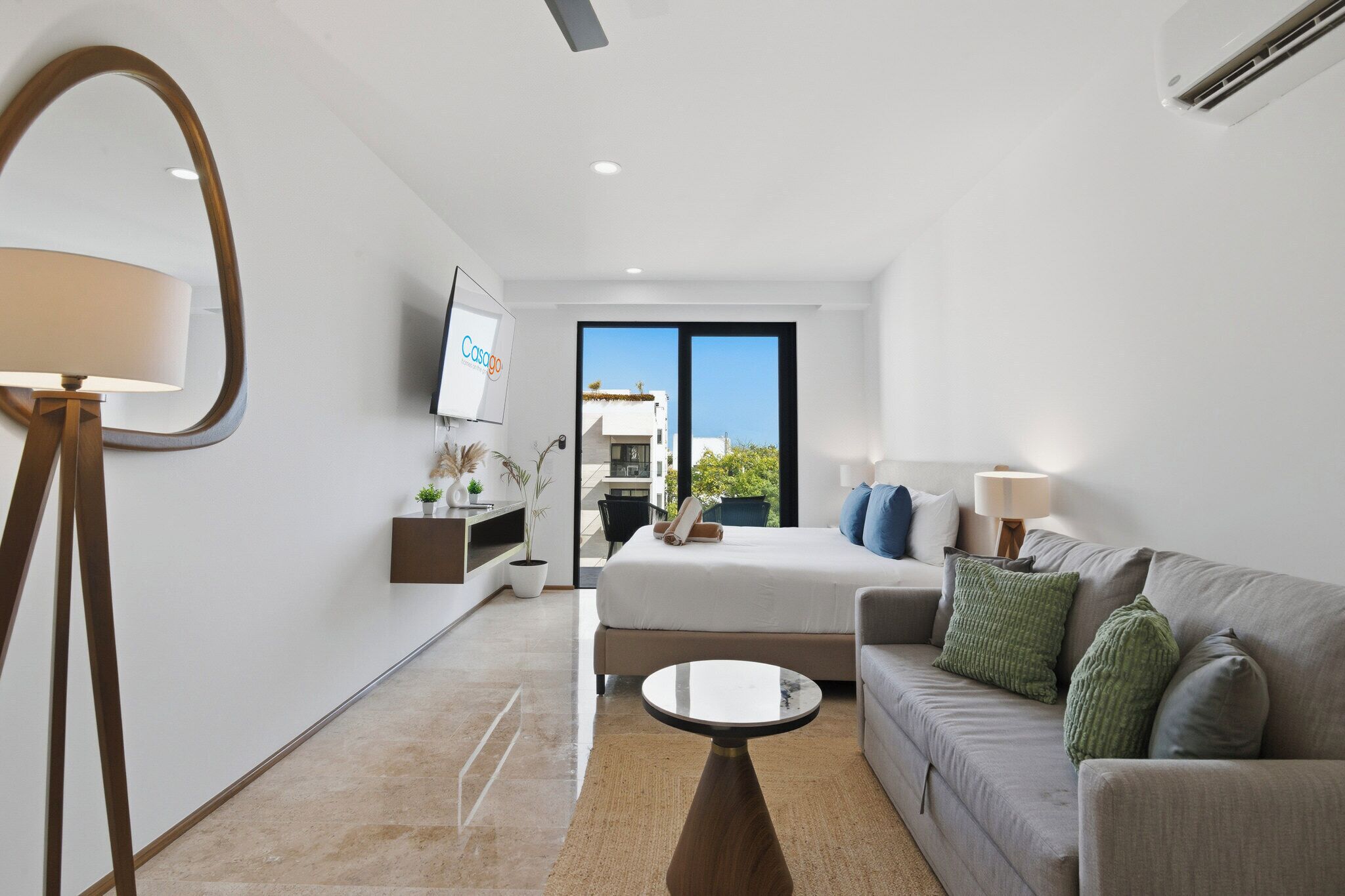 Habitación de hotel moderna con cama, sofá, mesa de centro, televisor y un amplio ventanal con vistas al exterior.