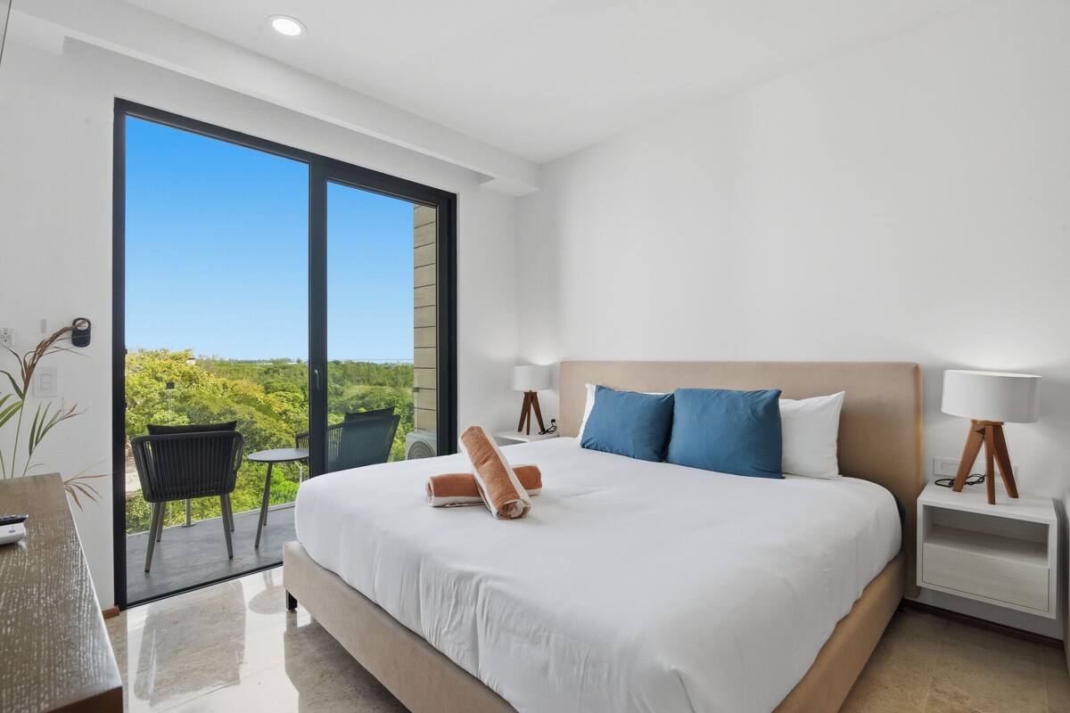 A modern hotel room with a large bed, a balcony with a view, a small table, and a chair.