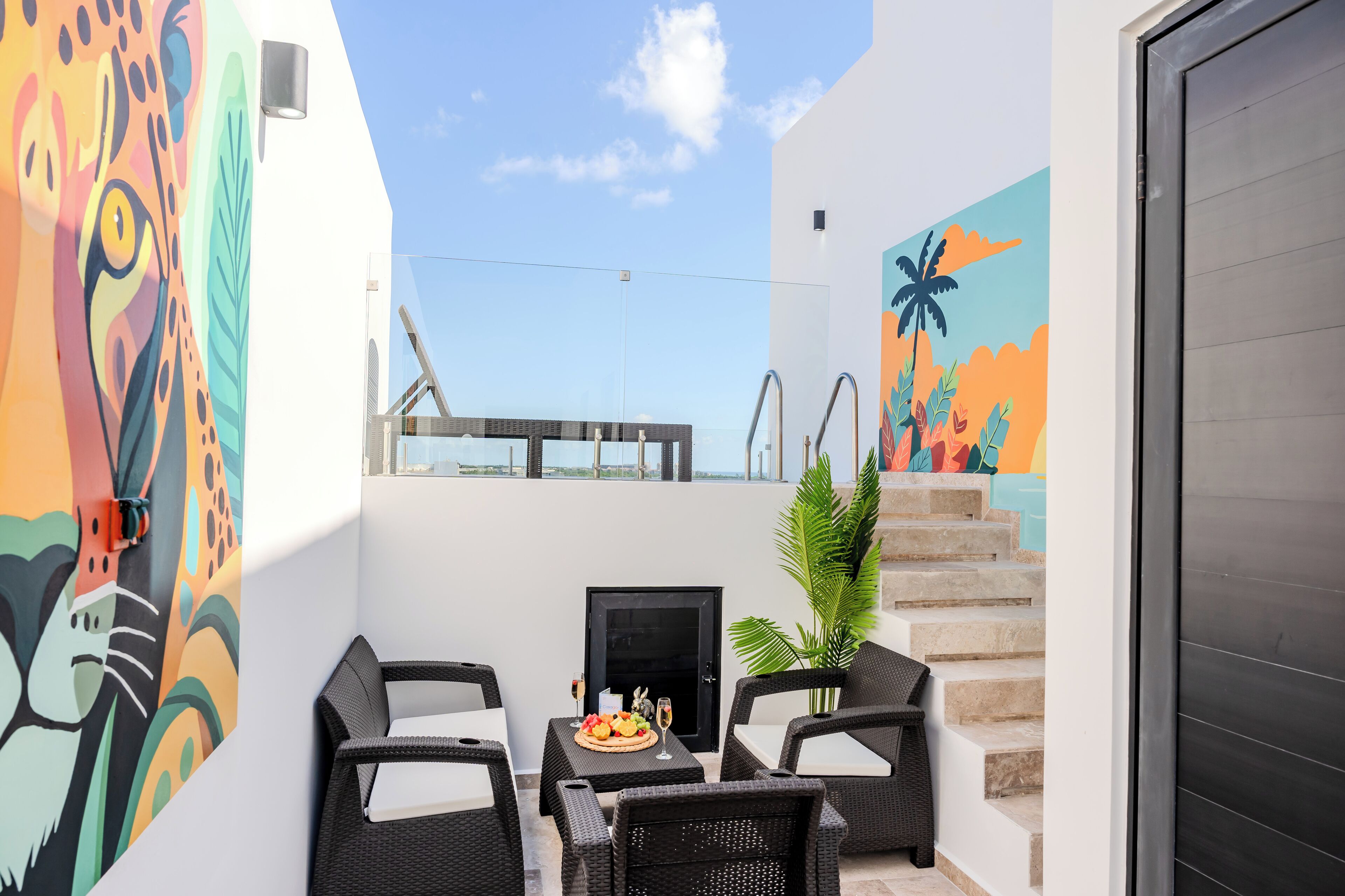 Eine Dachterrasse mit Korbmöbeln, einem kleinen Tisch mit Früchten und einem Glas sowie einem Wandgemälde.