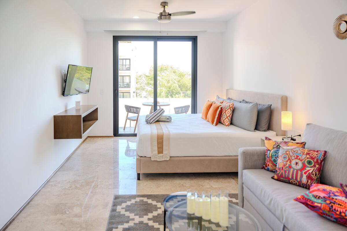 A modern hotel room with a bed, a sofa, a TV, a balcony, and a dining area.