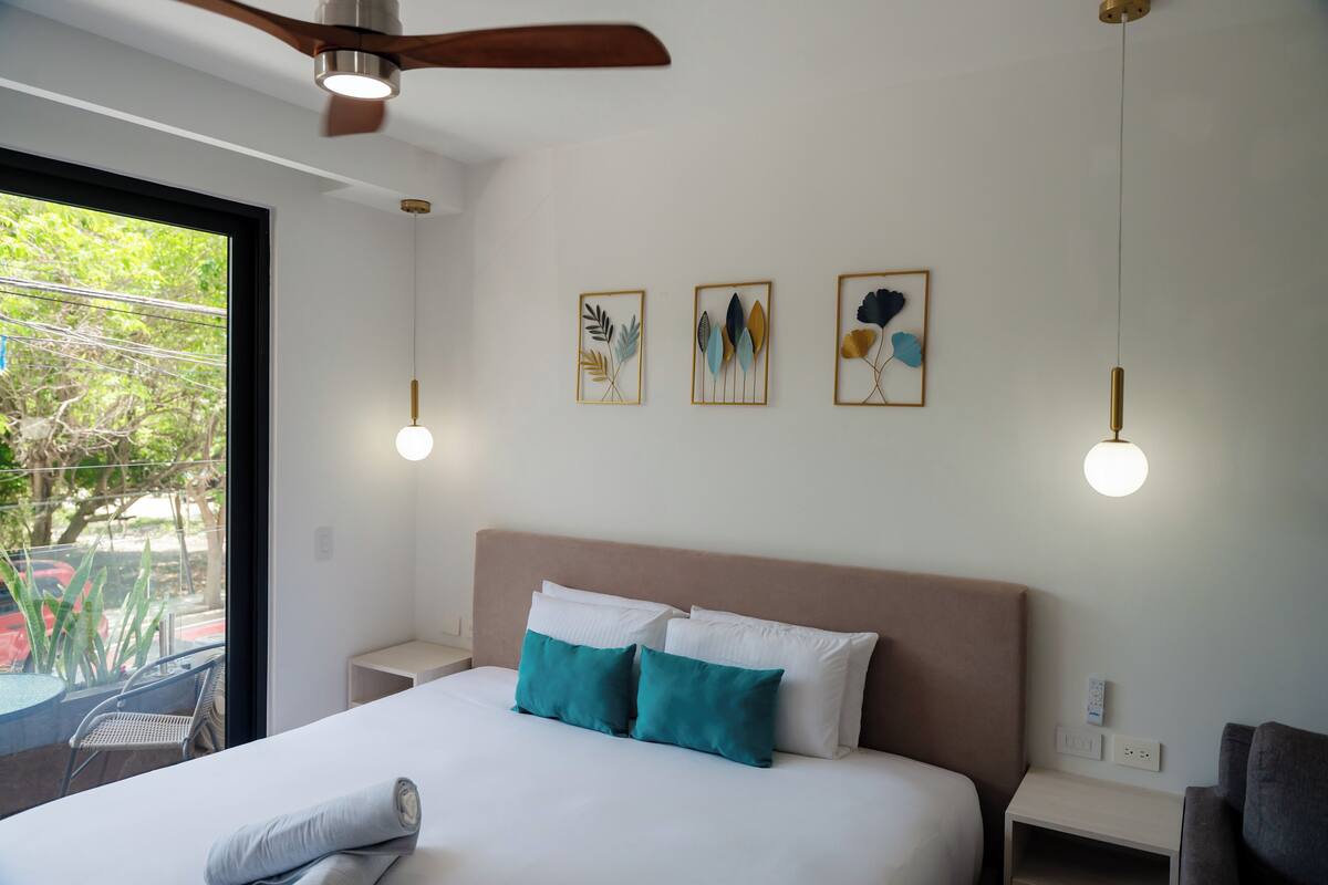 A hotel room with a bed, a ceiling fan, a window with a view of greenery, and wall-mounted artwork.