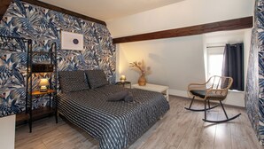 A bedroom with a bed, a nightstand, a lamp, a chair, and a wall with a tropical wallpaper.