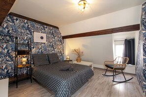 A bedroom with a bed, a nightstand, a lamp, a chair, and a wall with a tropical wallpaper.