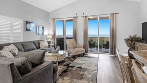 Un salón amplio con un sofá gris, un sillón estampado y un comedor con muebles de madera. Amplias ventanas ofrecen vistas al exterior.