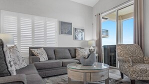 Una sala de estar con un sofá gris, un sillón estampado y una mesa de centro de vidrio. Hay dos lámparas de pared, un ventanal con persianas y un balcón con barandilla.