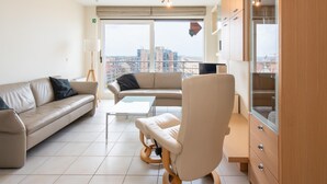 A modern living room with a sofa, armchair, and coffee table, large windows with a city view, and a wooden cabinet. - Apartment With Terrace and sea View in Mariakerke (Ostend)