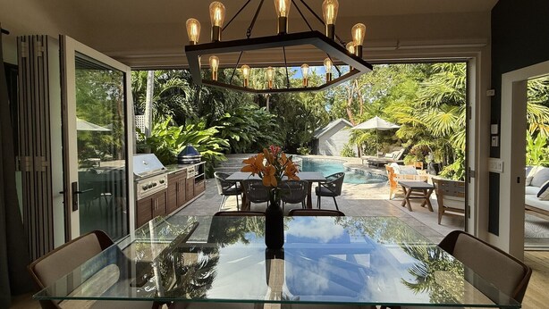 Un comedor moderno con una mesa y sillas de vidrio, una lámpara colgante y vistas a una piscina y jardín.