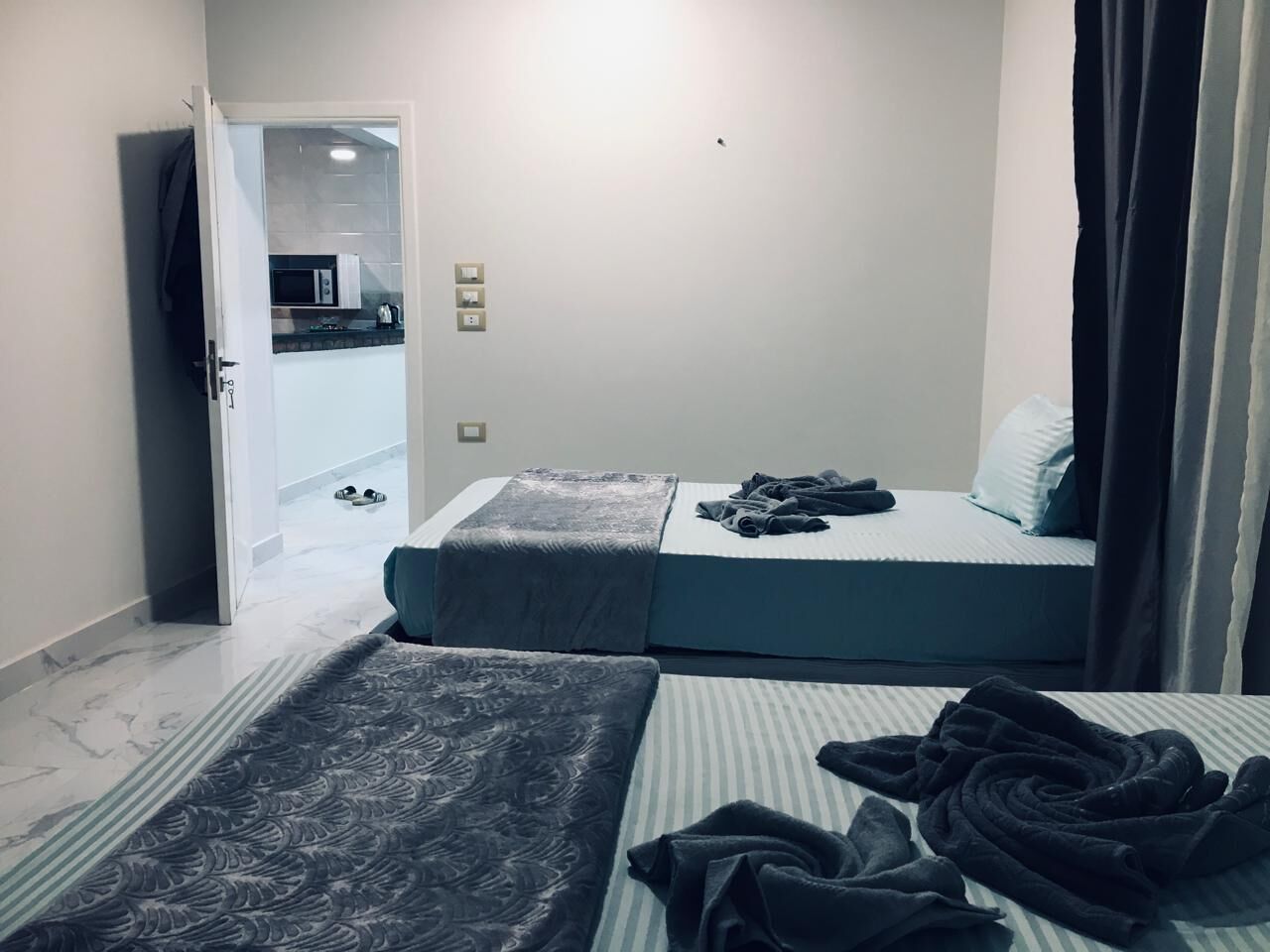 A small, compact bedroom with two beds, a visible kitchenette in the background, and a door leading to another room.
