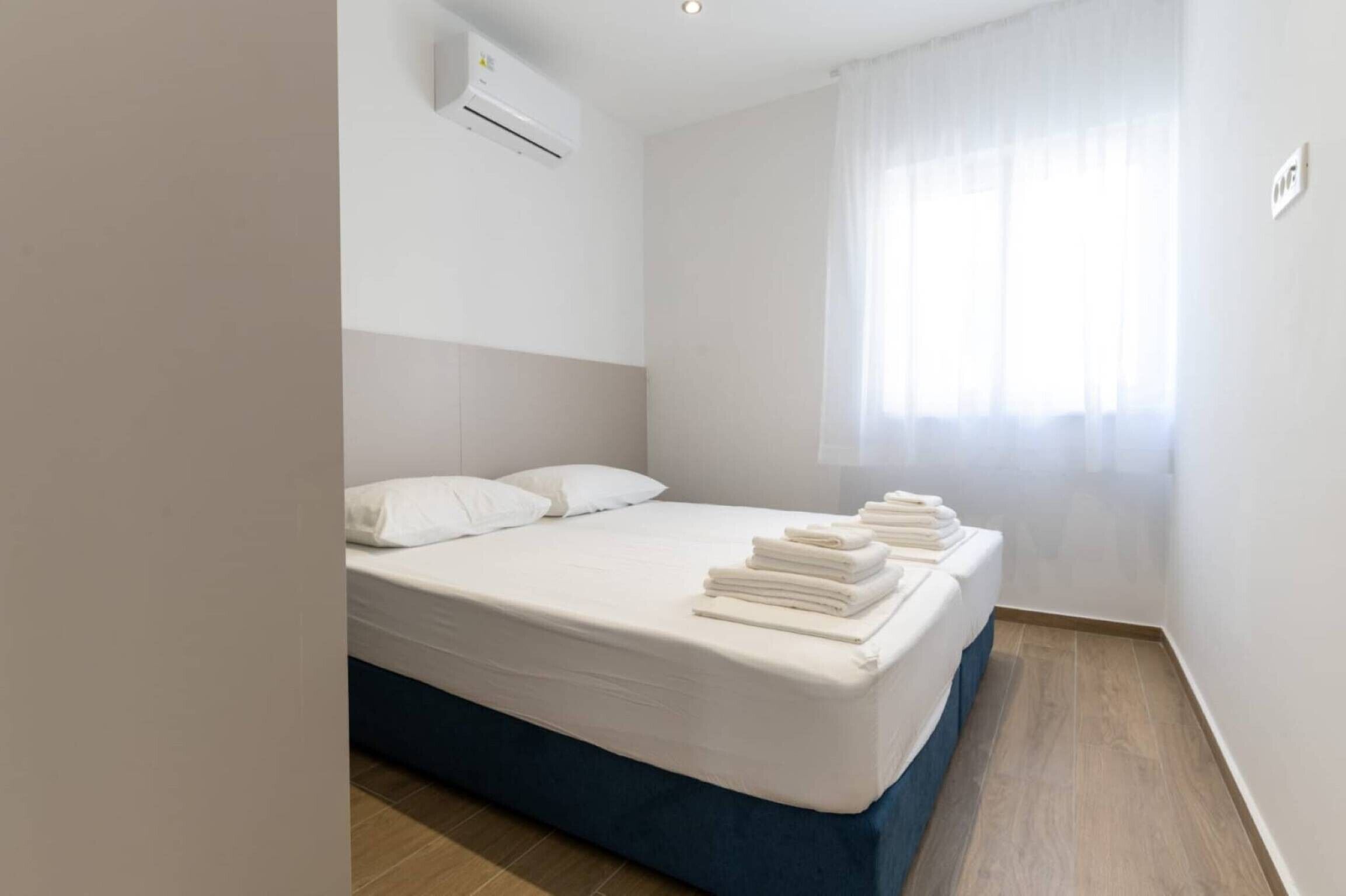A hotel room with a bed, white linens, a blue mattress cover, a window with sheer curtains, and an air conditioning unit on the wall.