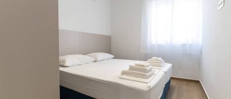 A hotel room with a bed, white linens, a blue mattress cover, a window with sheer curtains, and an air conditioning unit on the wall.