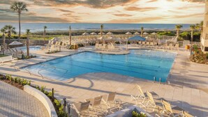 A swimming pool with lounge chairs and umbrellas, surrounded by palm trees and a view of the ocean. - MyStay at 1208 Villamare • Ocean views • Premier Design • New Pools & Hot Tubs! (Hilton Head Island)