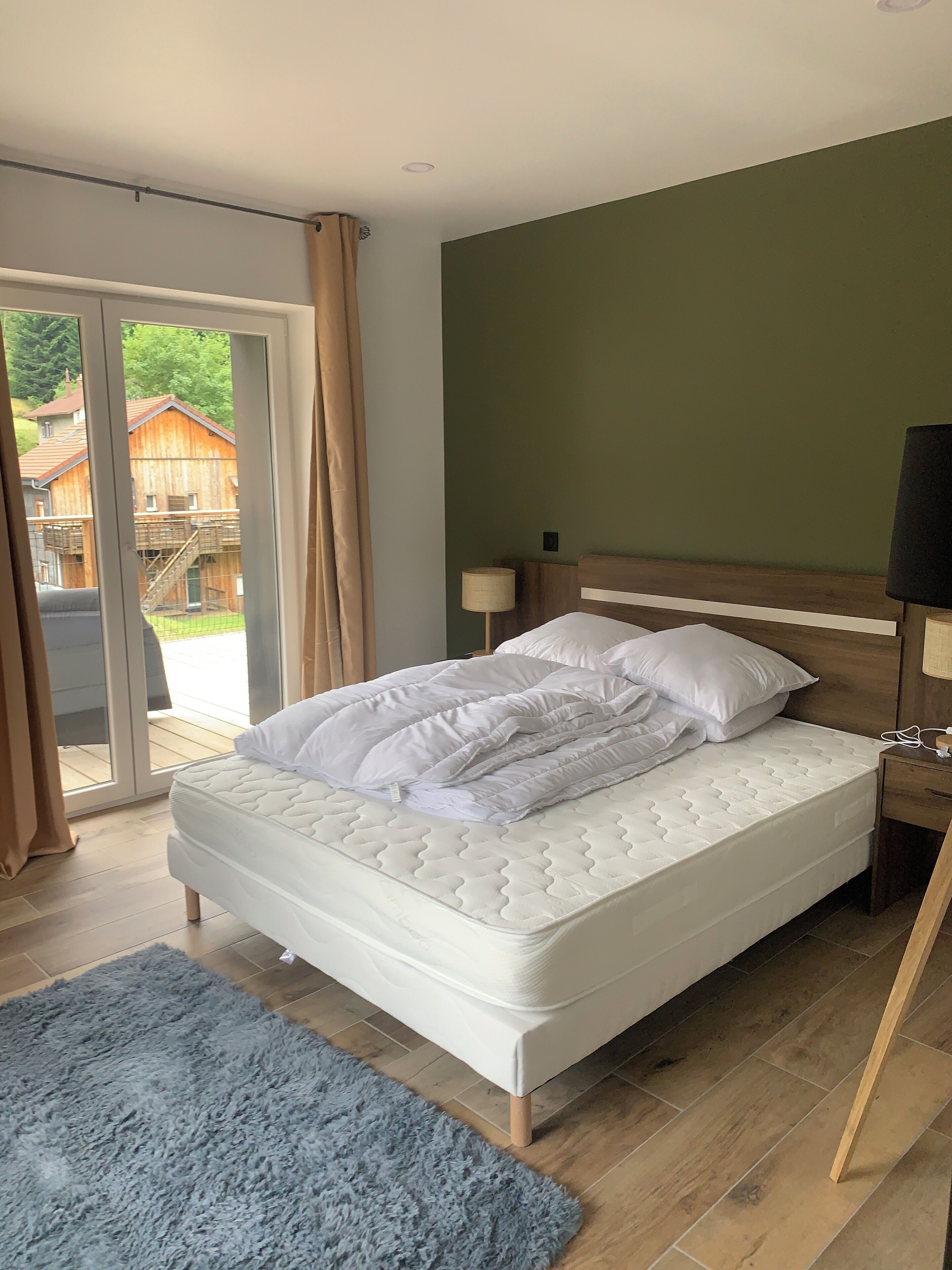 Een slaapkamer met een groot bed, een houten hoofdbord, een nachtkastje met een lamp en uitzicht op een houten gebouw door een glazen deur.