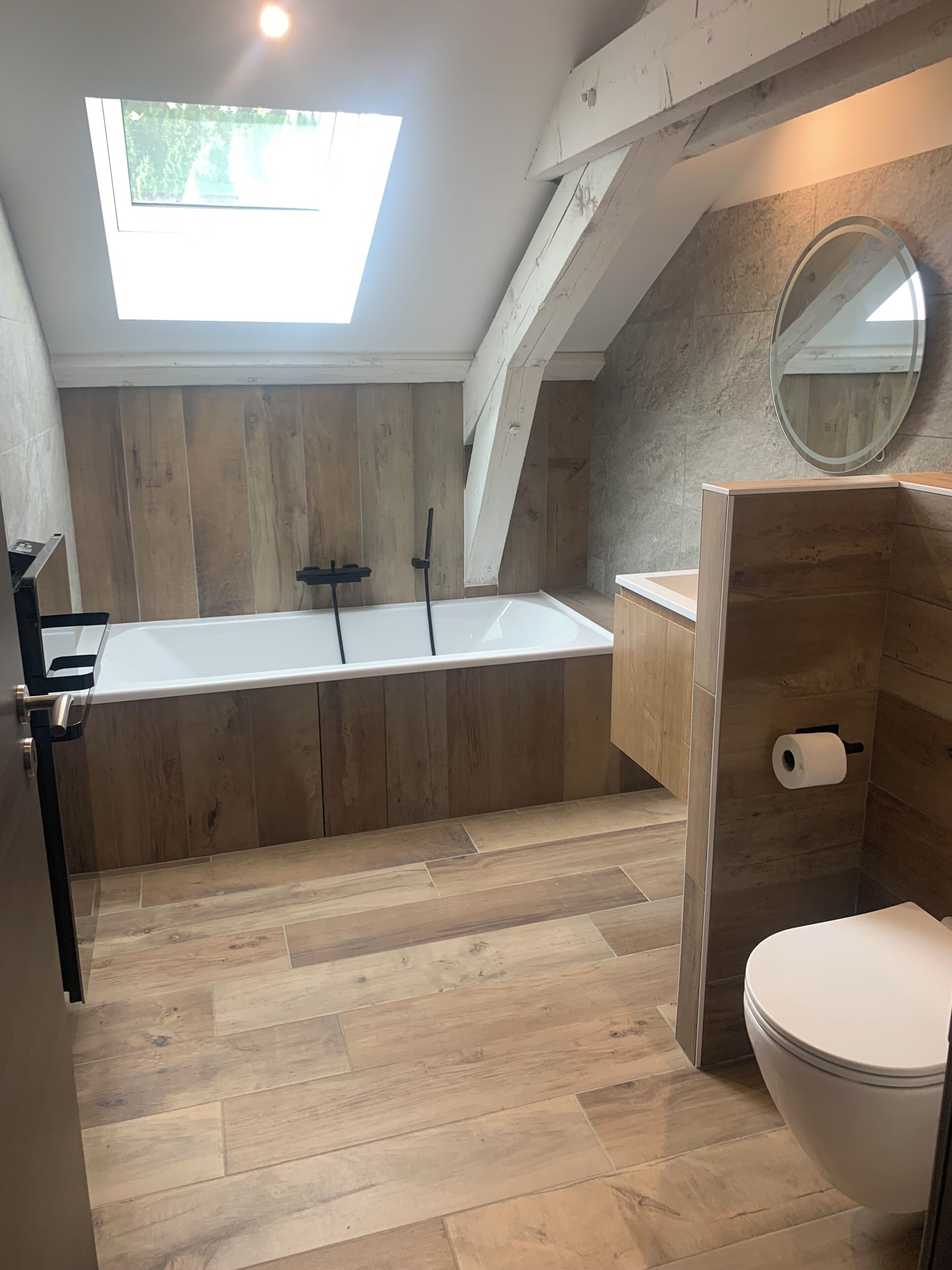 Een moderne badkamer met dakraam, houten vloer, een vrijstaand bad, toilet en spiegel.