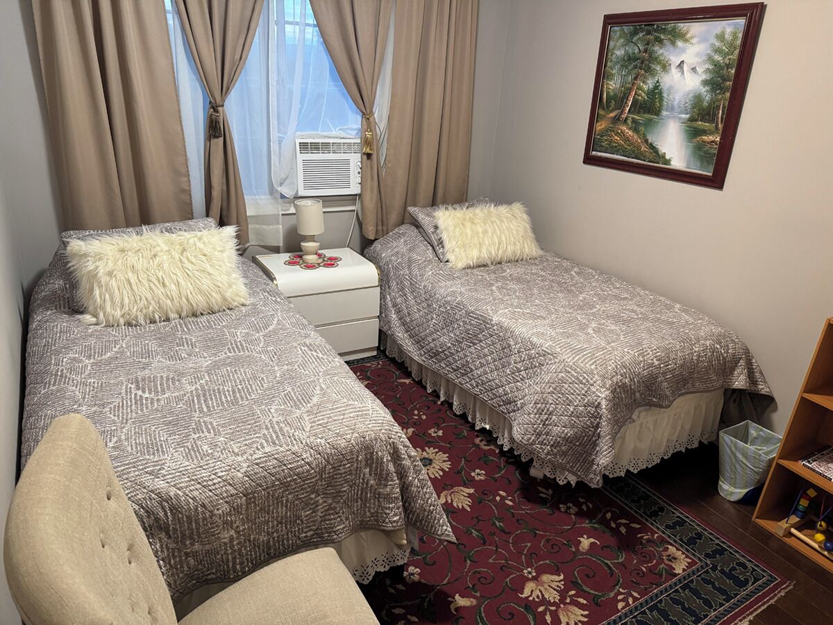 A bedroom with two beds, a nightstand, a lamp, a window with curtains, a painting, and a bookshelf.