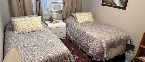 A bedroom with two beds, a nightstand, a lamp, a window with curtains, a painting, and a bookshelf.