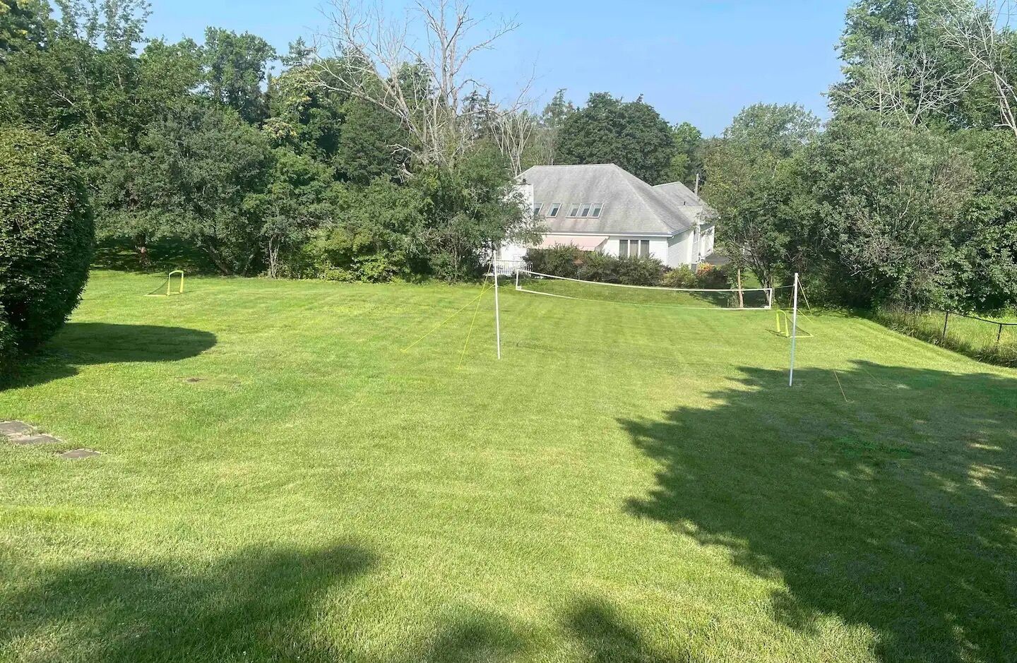 Une pelouse bien entretenue avec un filet de volley-ball, entourée d’arbres et avec une maison en arrière-plan.